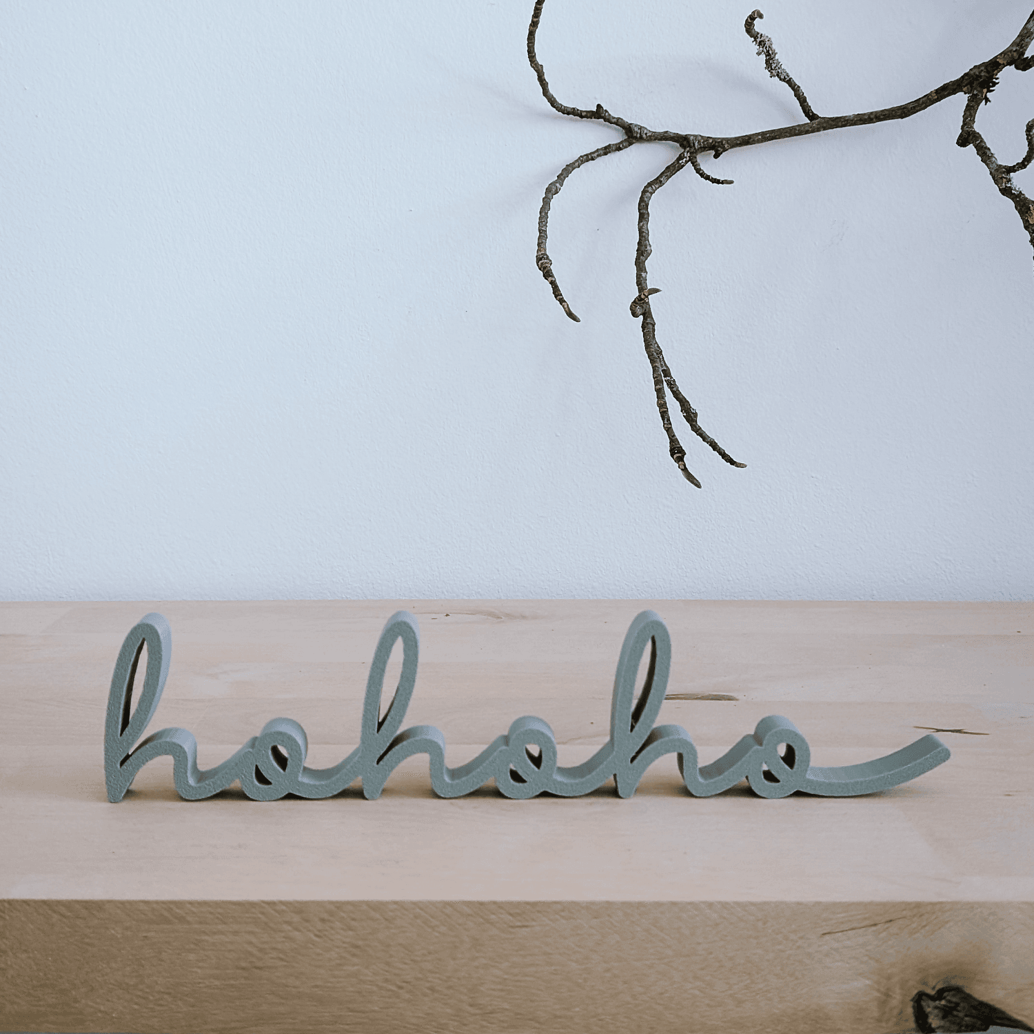 'hohoho' Schriftzug in Salbeigrün auf Holztisch vor weißer Wand mit Zweig